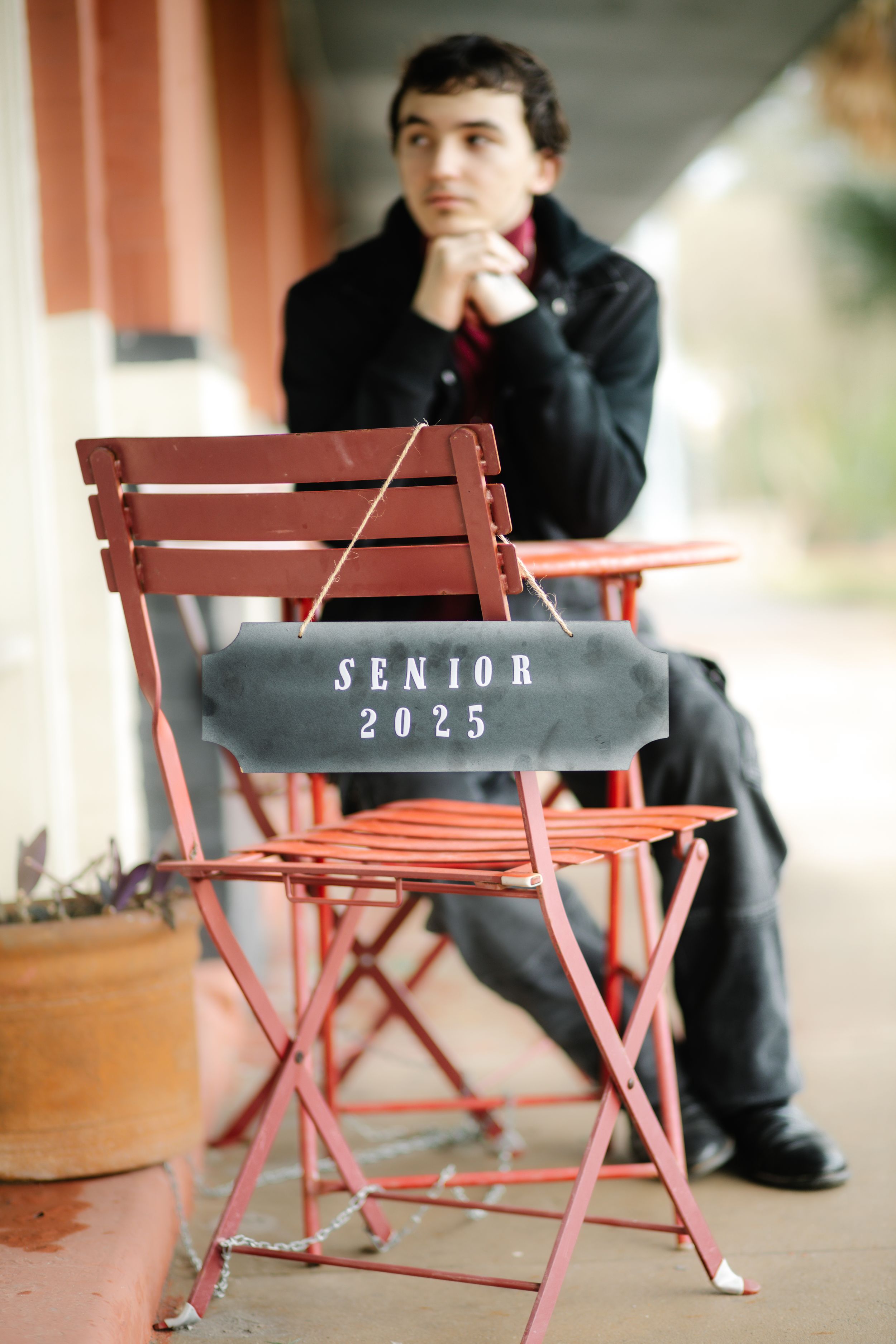 A red folding chair with 'Senior 2025' text sits in front of someone in formal attire in a thoughtful pose.