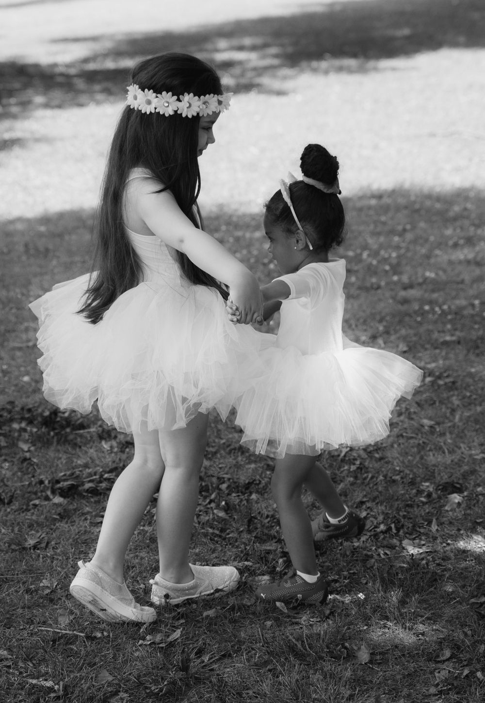 Two young dancers in white tutus hold hands and twirl together in a graceful black and white moment.
