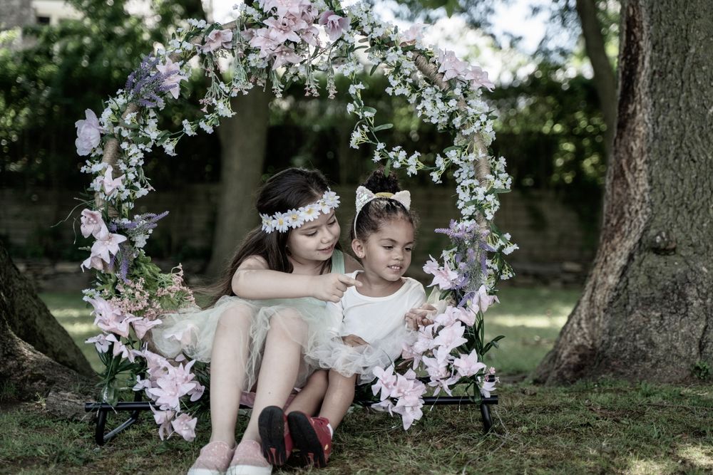 Two children sit together on a floral arch swing in a dreamy garden setting.