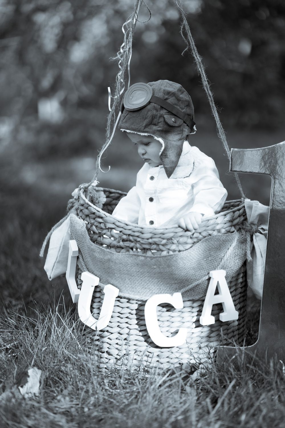 A child wearing aviator goggles sits in a decorative basket labeled USA in a whimsical black and white photo.