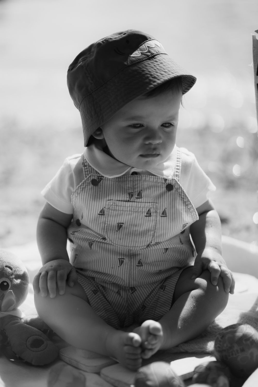 A baby in overalls and baseball cap sits outdoors in a vintage-style black and white portrait.