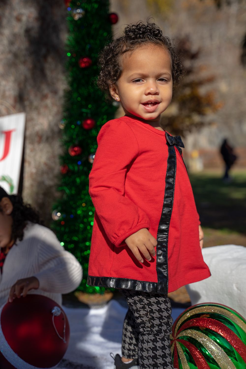 A small child in a red top and patterned pants poses in front of a decorated Christmas tree outdoors.
