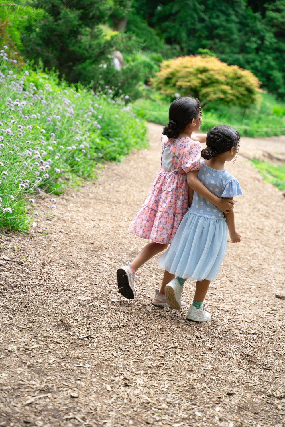 Two children in summer dresses walk hand in hand along a garden path surrounded by blooming flowers.