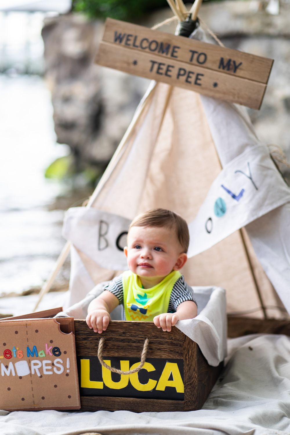 Baby sits in wooden box labeled 'Luca' next to teepee tent with welcome sign.