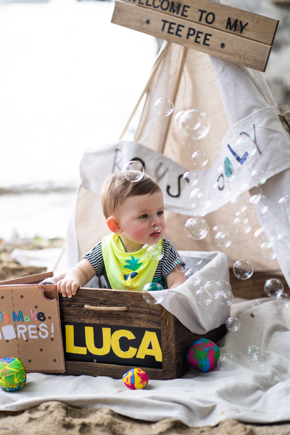 Baby plays with bubbles in wooden box near teepee tent with 'Welcome' sign.