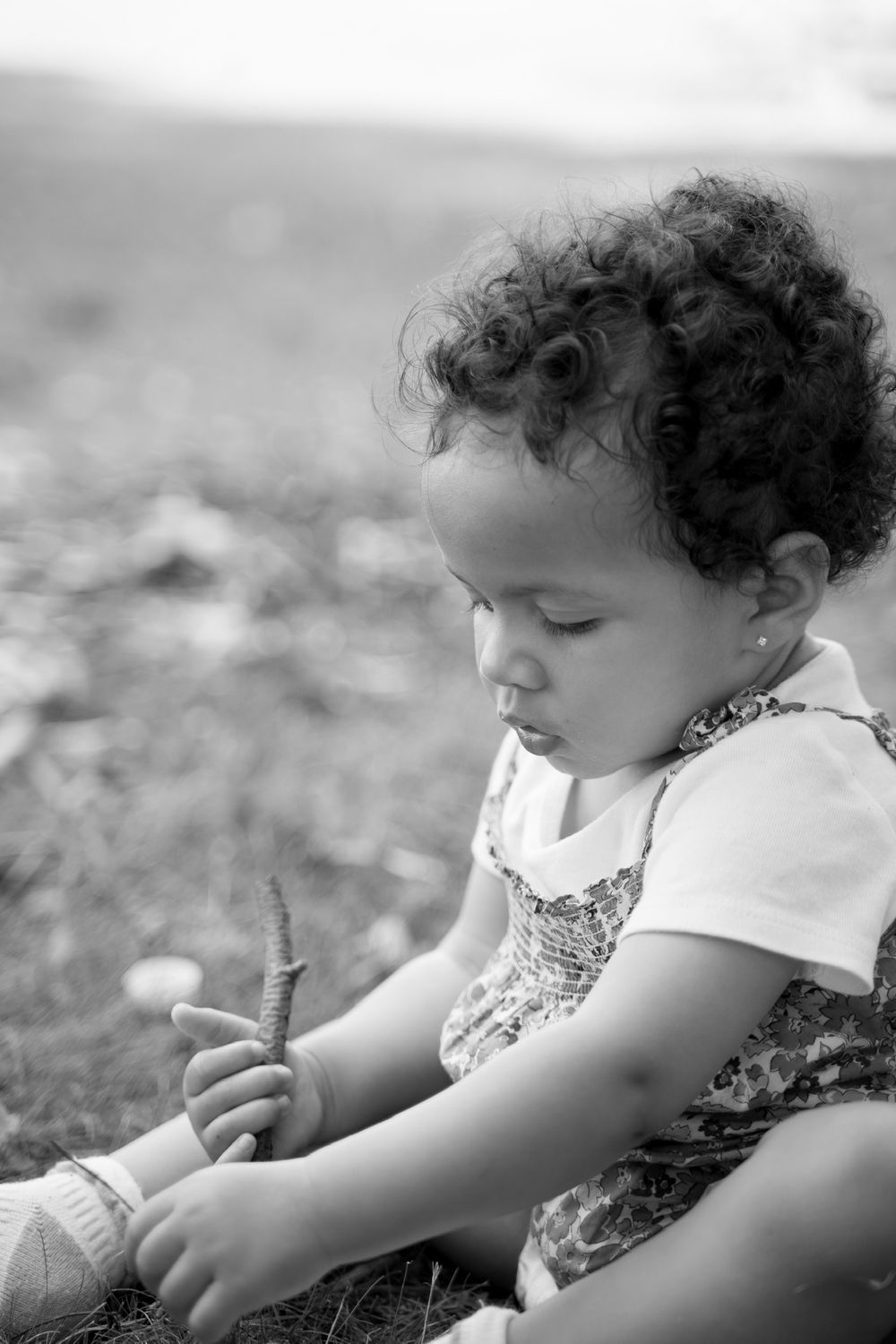 A small child with curly hair explores something interesting in the grass while sitting outdoors in black and white.