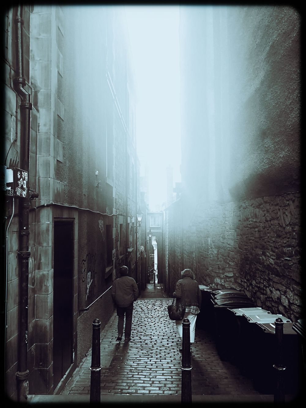 Foggy cobblestone alley at night with dark silhouettes creating an eerie atmospheric scene in black and white.