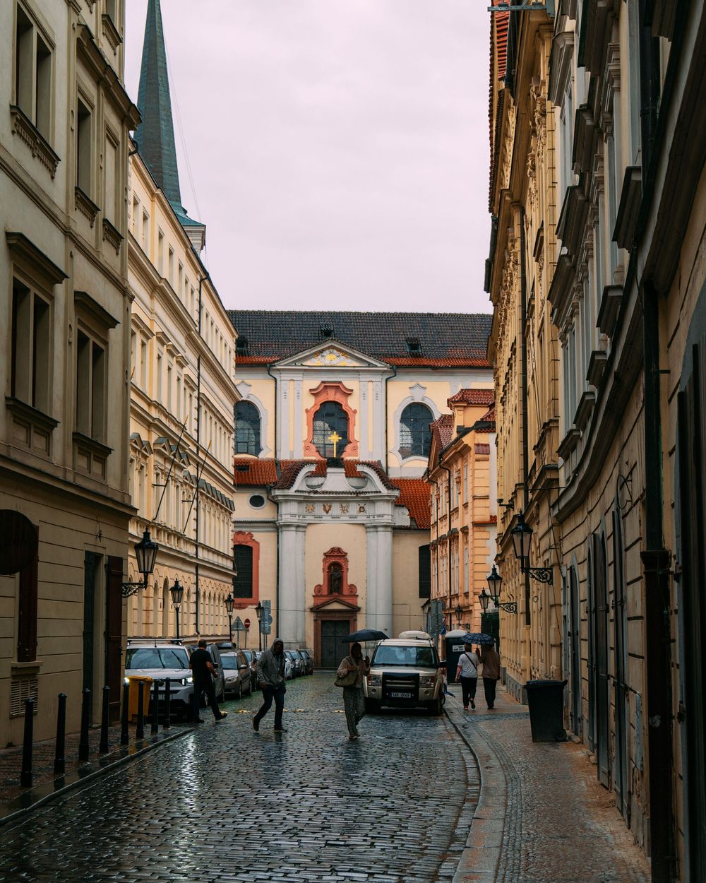 Historic cobblestone street in Prague leads to a church with baroque architecture and arched windows.