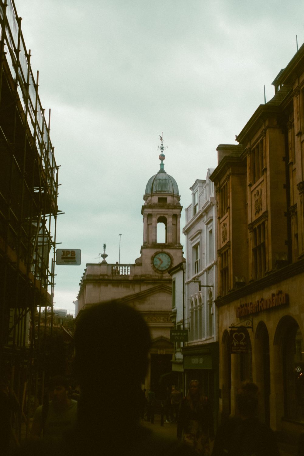 Historic church tower with domed spire rises above a busy shopping street with scaffolding in moody overcast weather.
