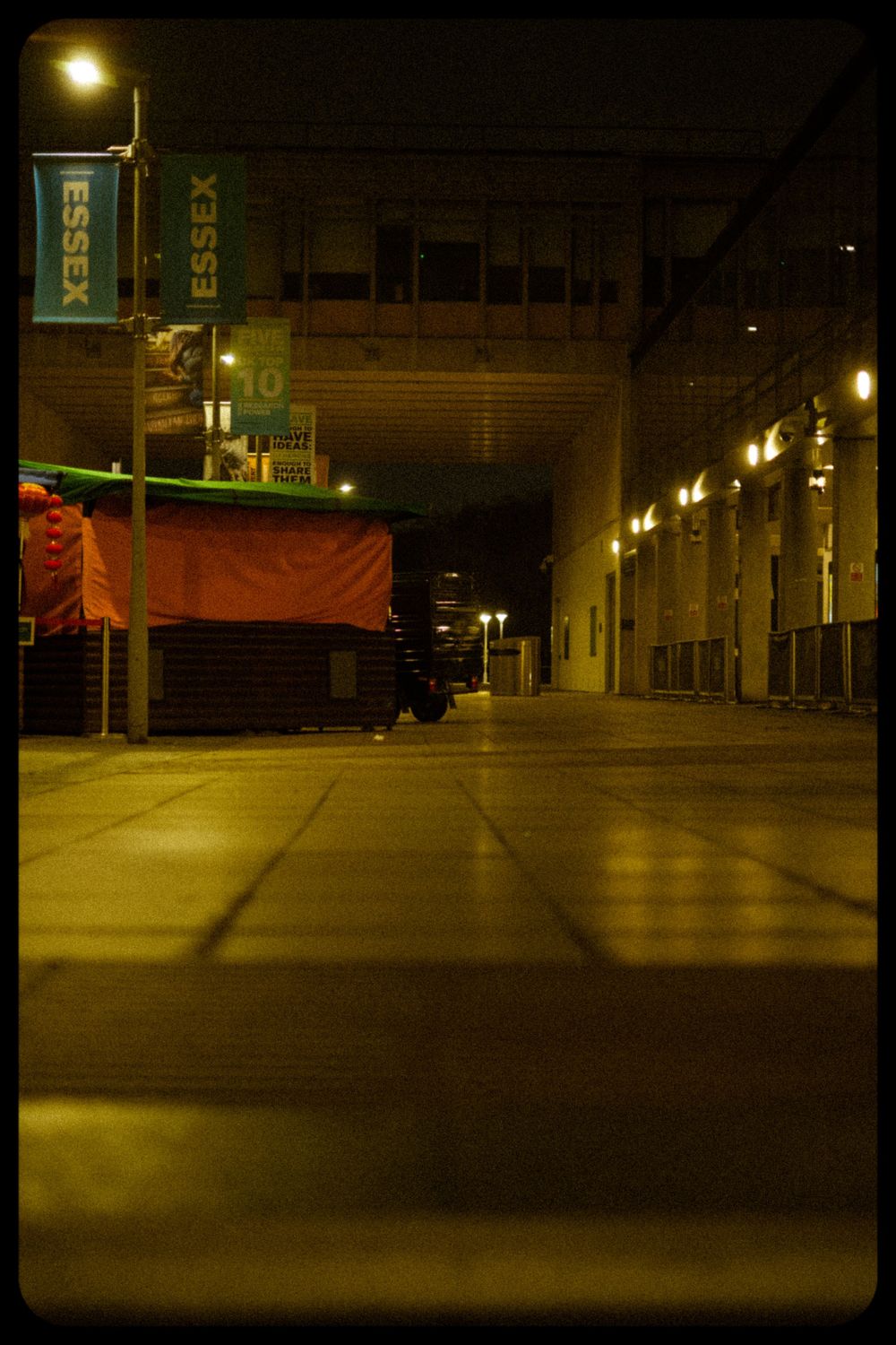 A dimly lit urban street at night with illuminated lampposts and Essex banners along a concrete building facade.