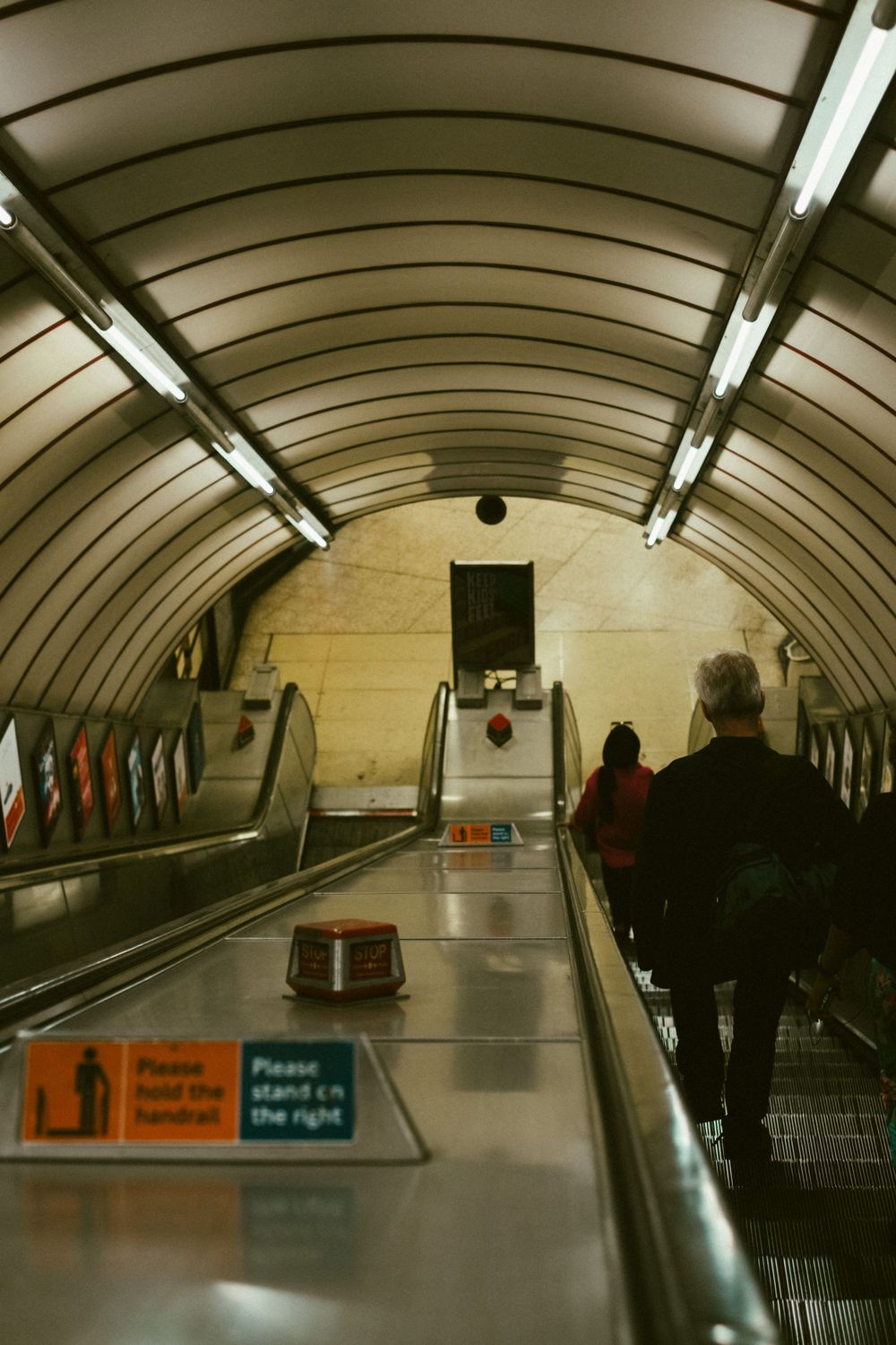 Curved escalator tunnel in London Underground station with fluorescent lighting and directional signs mounted on wall.