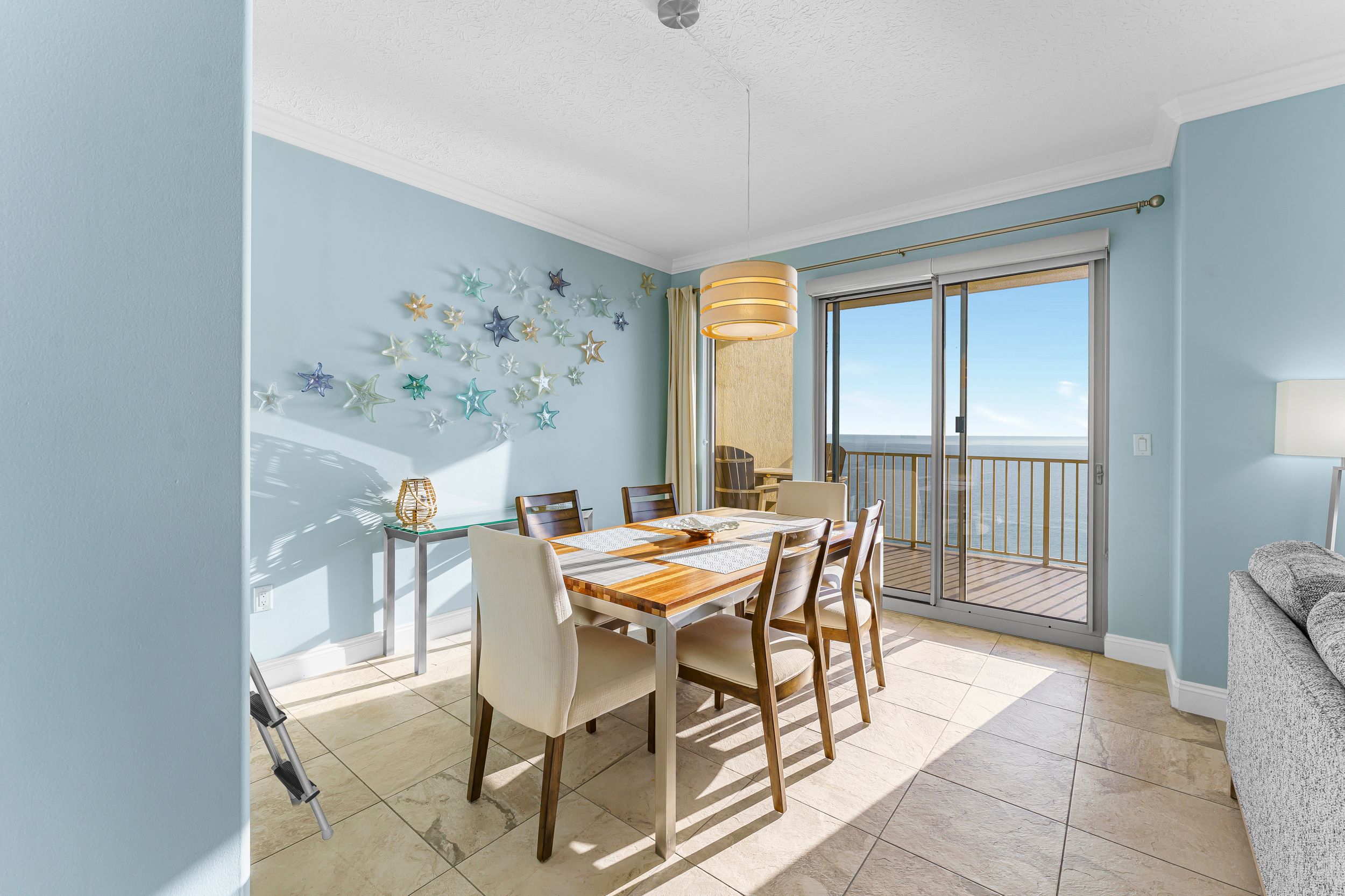 Bright coastal dining area with light blue walls, wooden table, and balcony access overlooking the beach.