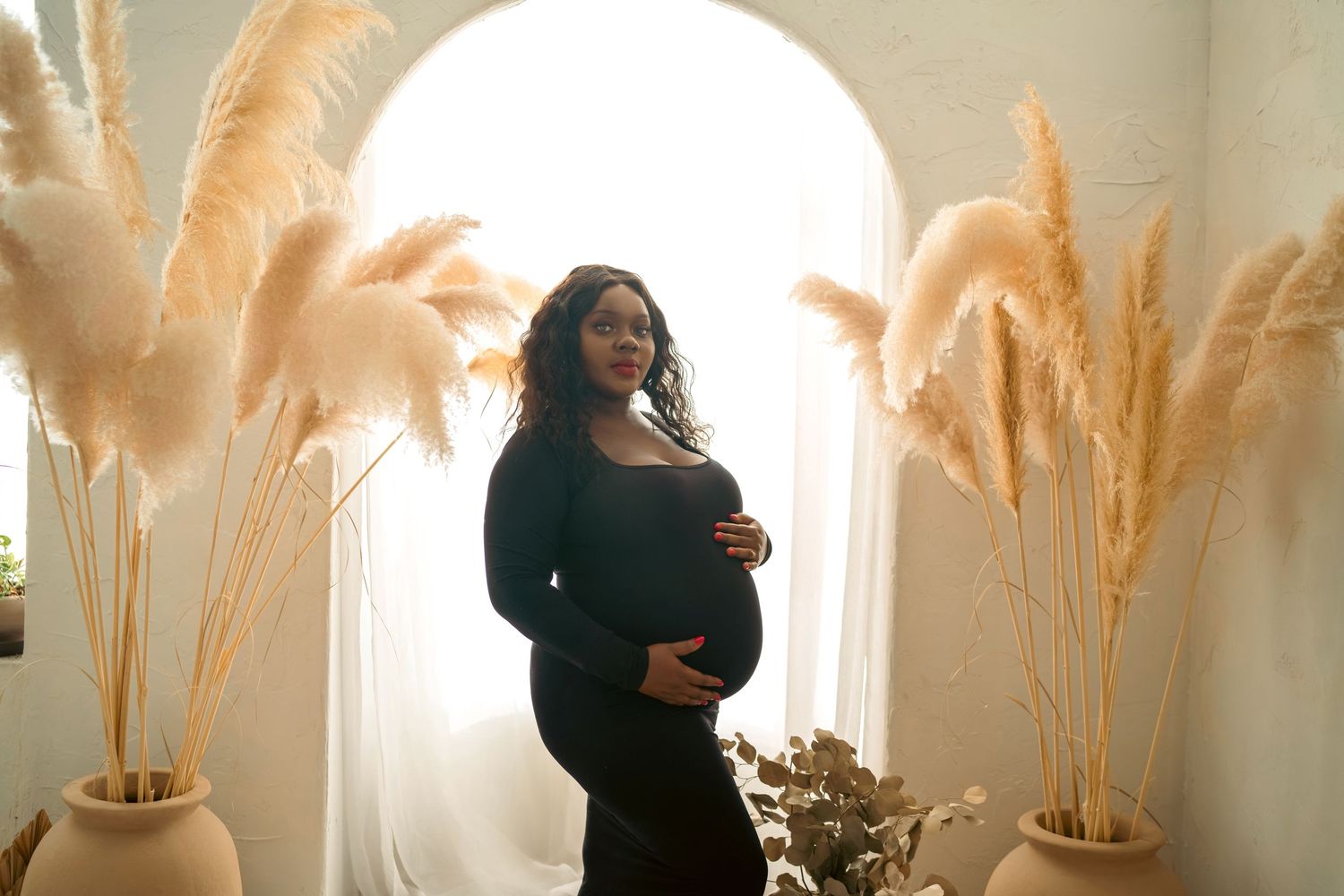 Dramatic maternity silhouette against arched window with dried pampas grass decor.