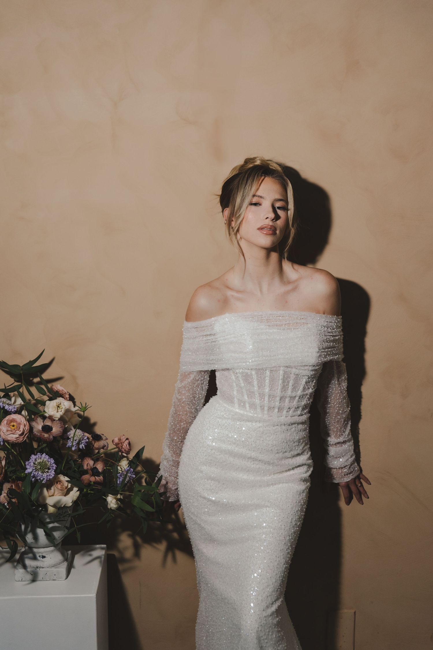 An elegant off-shoulder white sequined wedding gown is styled with a floral arrangement against a neutral wall.