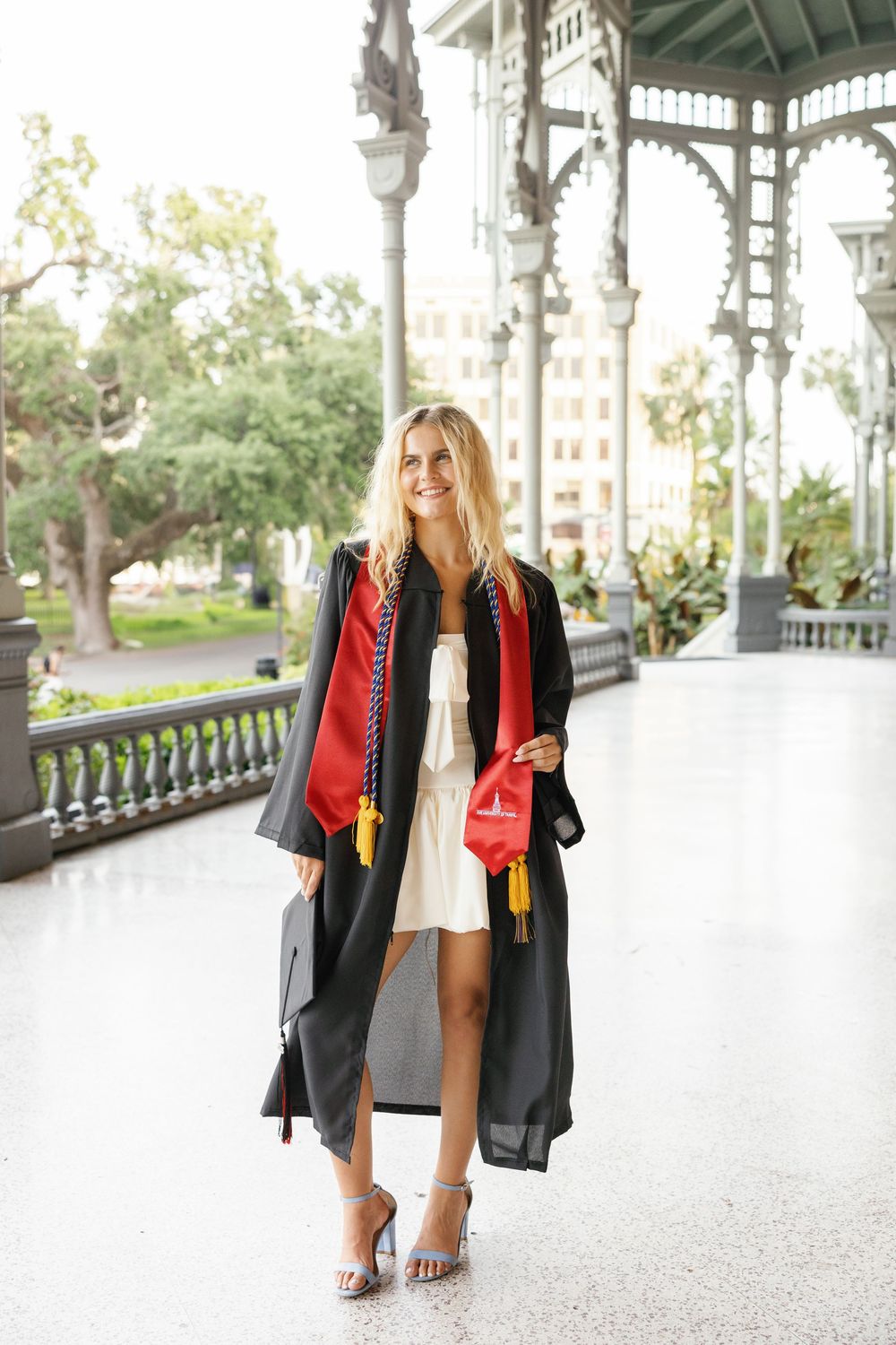 Graduate wearing academic regalia with red and black graduation gown stands smiling on ornate white porch.