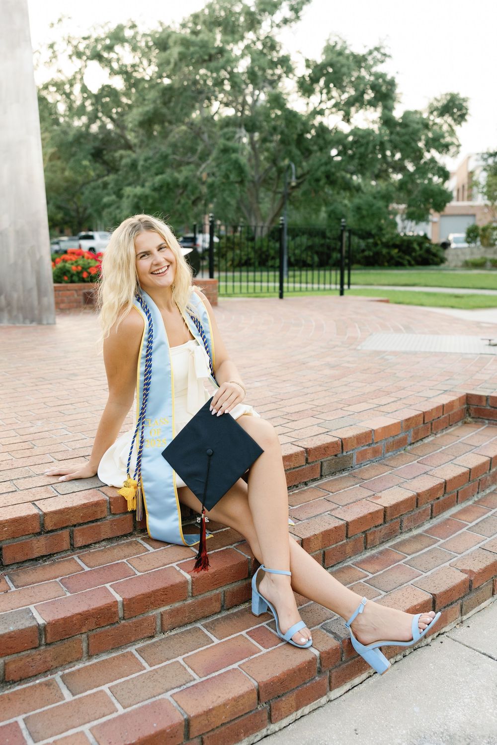 Graduate in a white dress sitting on brick steps smiling while holding a mortarboard graduation cap.