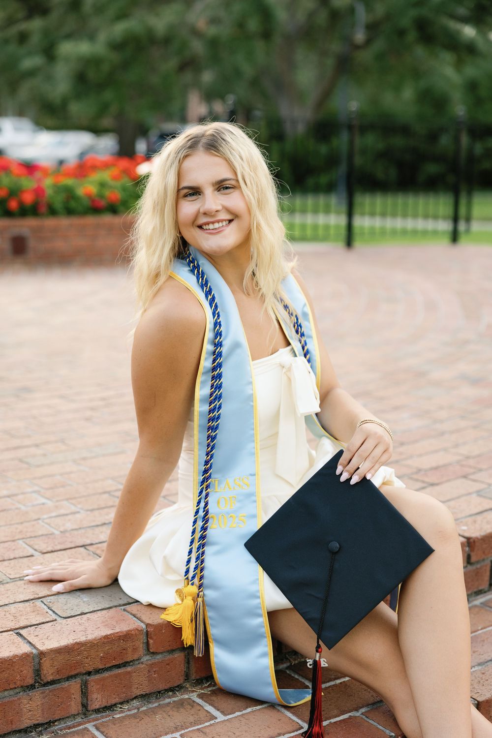 Graduate wearing a white dress and blue graduation stole sits on brick wall with mortarboard cap in outdoor setting.