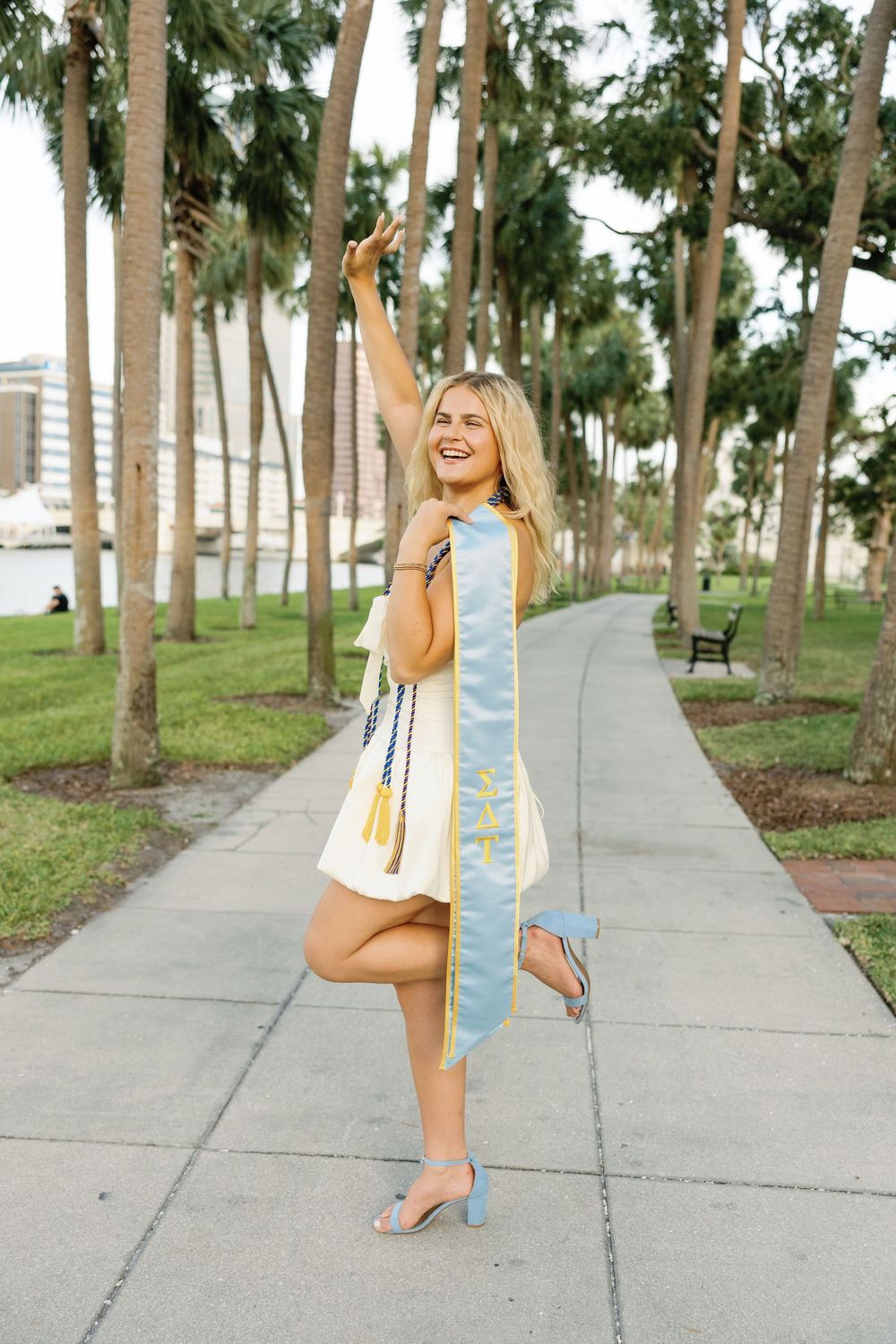 Graduate celebrating in a blue graduation stole and white dress poses on palm tree-lined walkway.