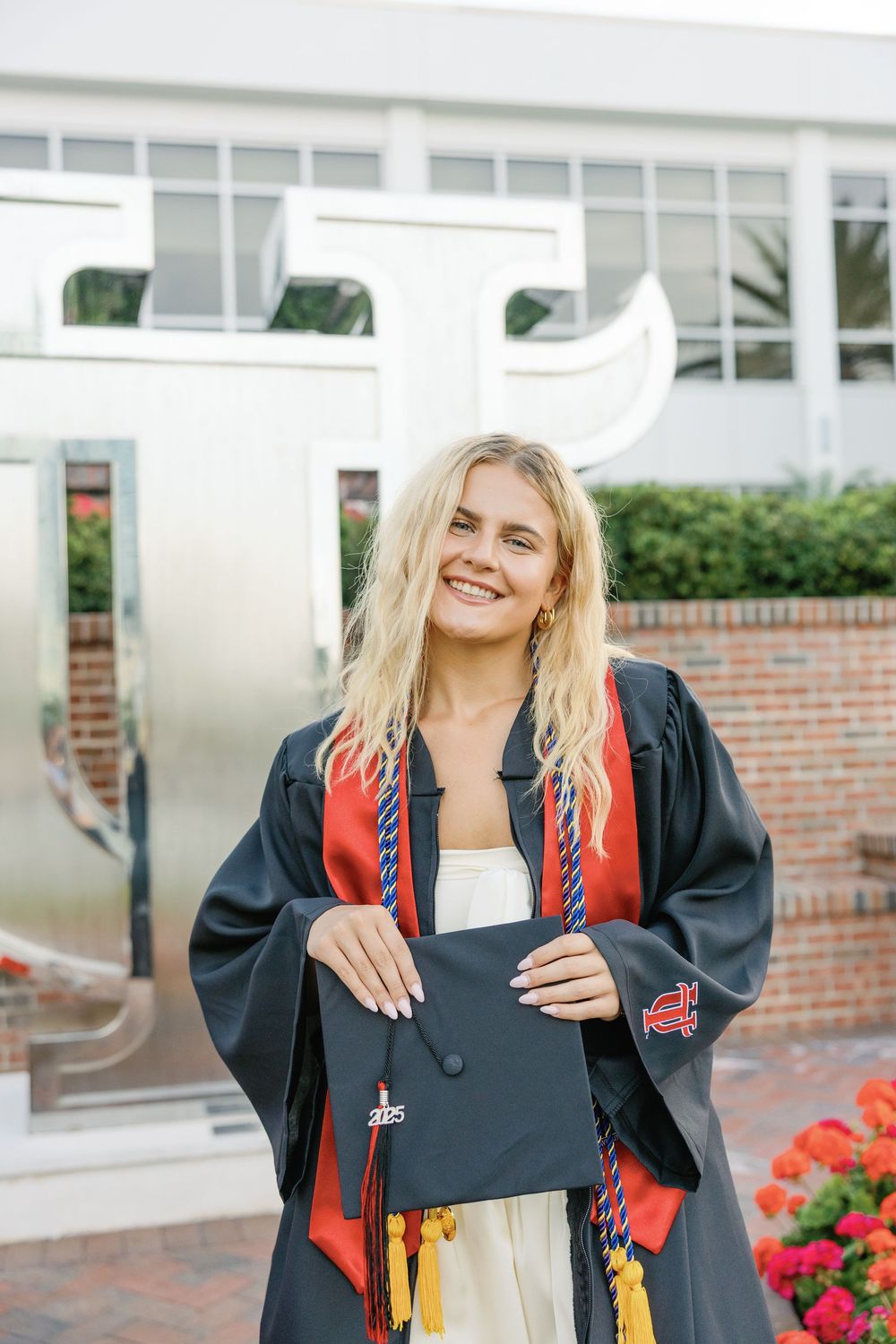 Graduate in black cap and gown holds decorated graduation cap while standing outdoors by brick building with flowers.
