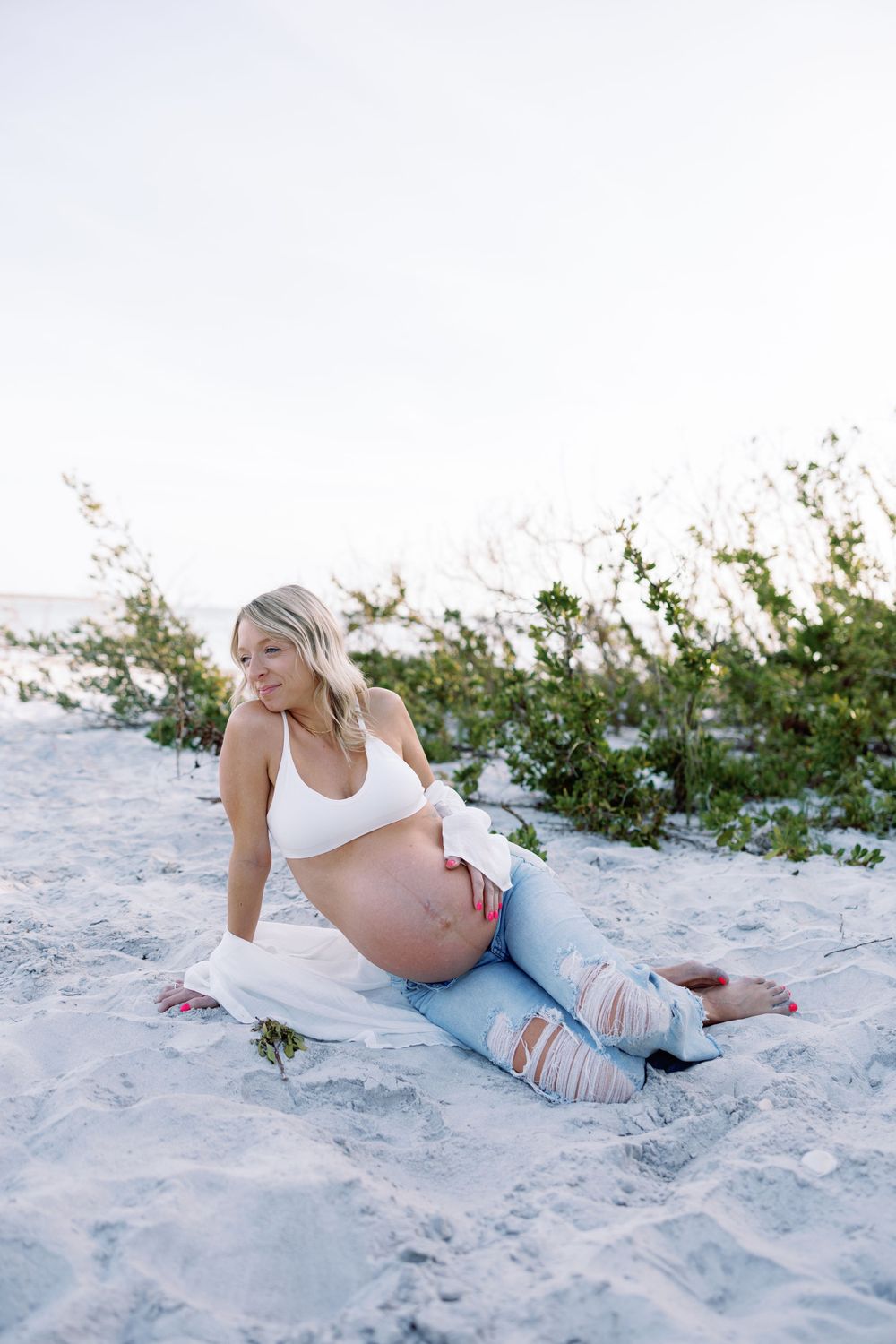 A pregnant woman in distressed jeans and white top reclines on a sandy beach at sunset.