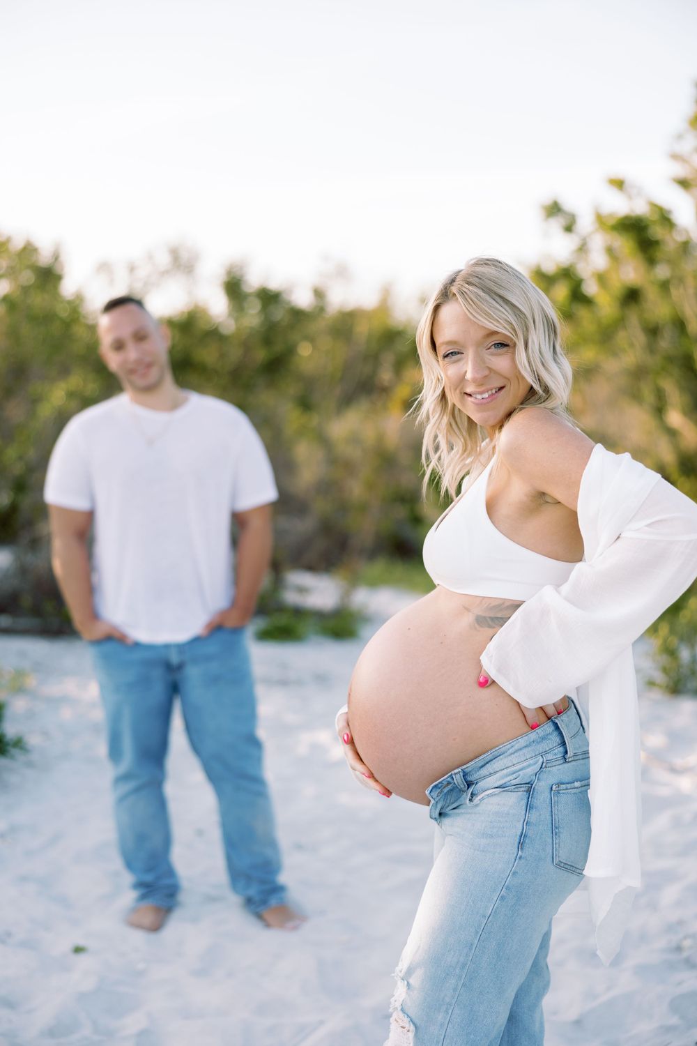 Expecting couple poses for maternity photos on a sandy beach with soft natural lighting at sunset.