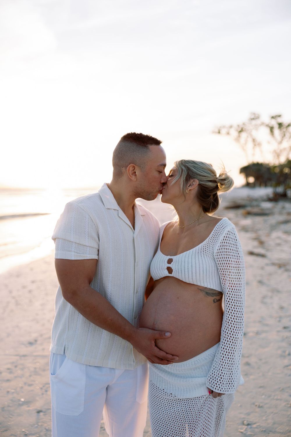 A couple shares an intimate moment on a beach at sunset, embracing a pregnant belly in matching white outfits.