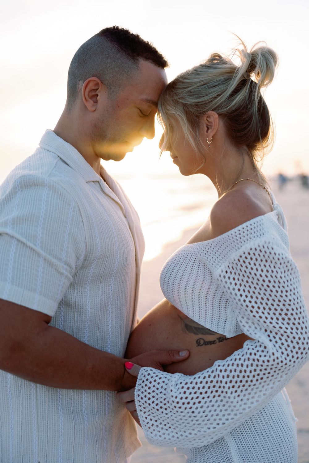 An intimate maternity photo of a couple touching foreheads at sunset, with the woman wearing a white knit sweater.