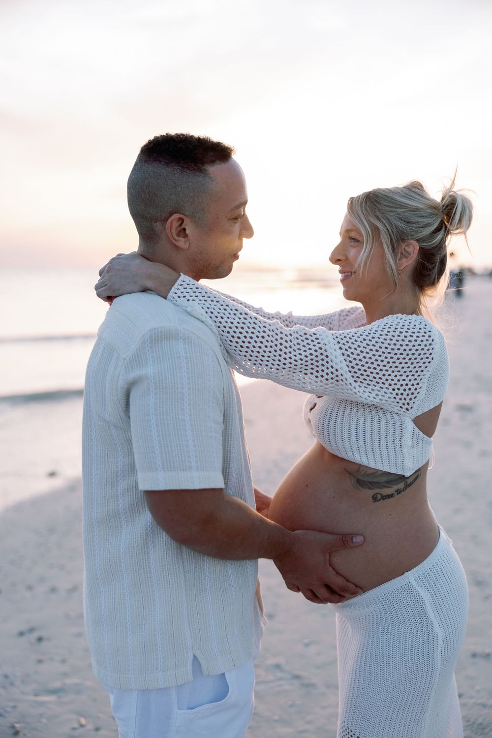 A romantic couple embraces on a beach at sunset, with the pregnant woman wearing a white knit outfit.
