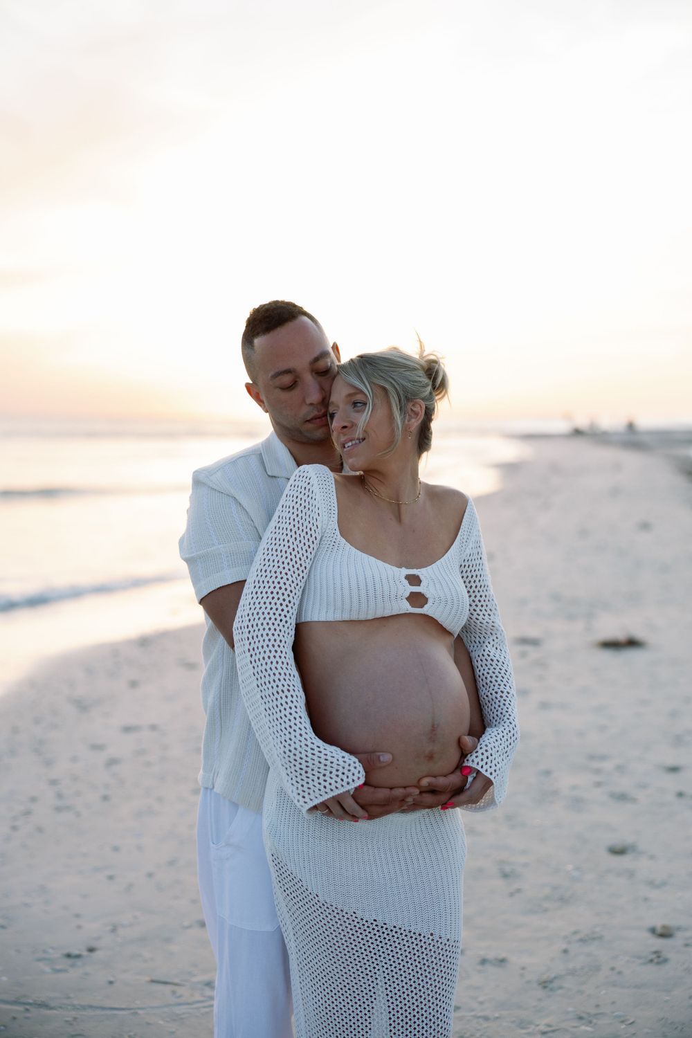 An expectant couple in white attire embraces on a tranquil beach during golden hour.