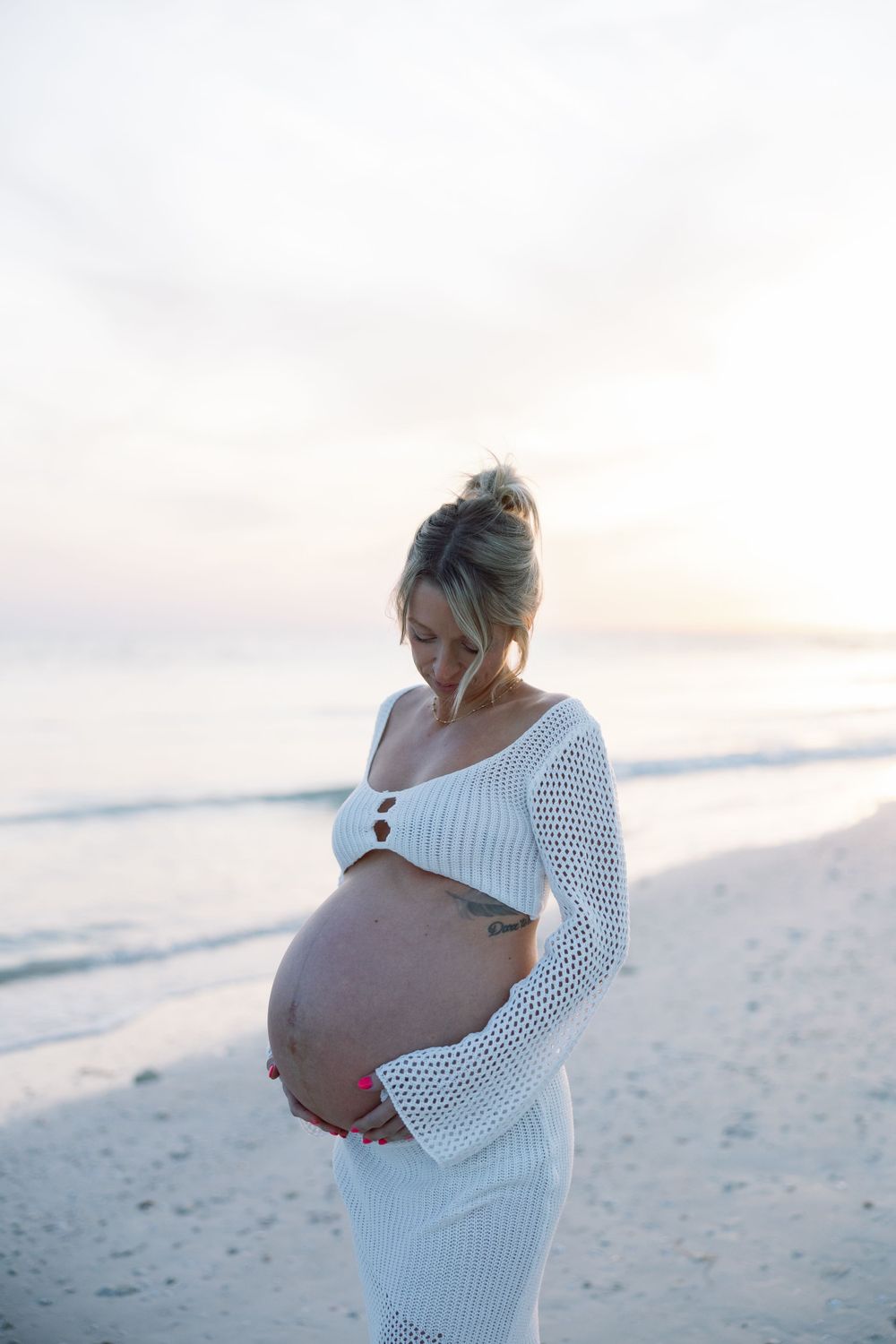 An expectant mother in a white knit dress stands on a beach during sunset, cradling her baby bump.