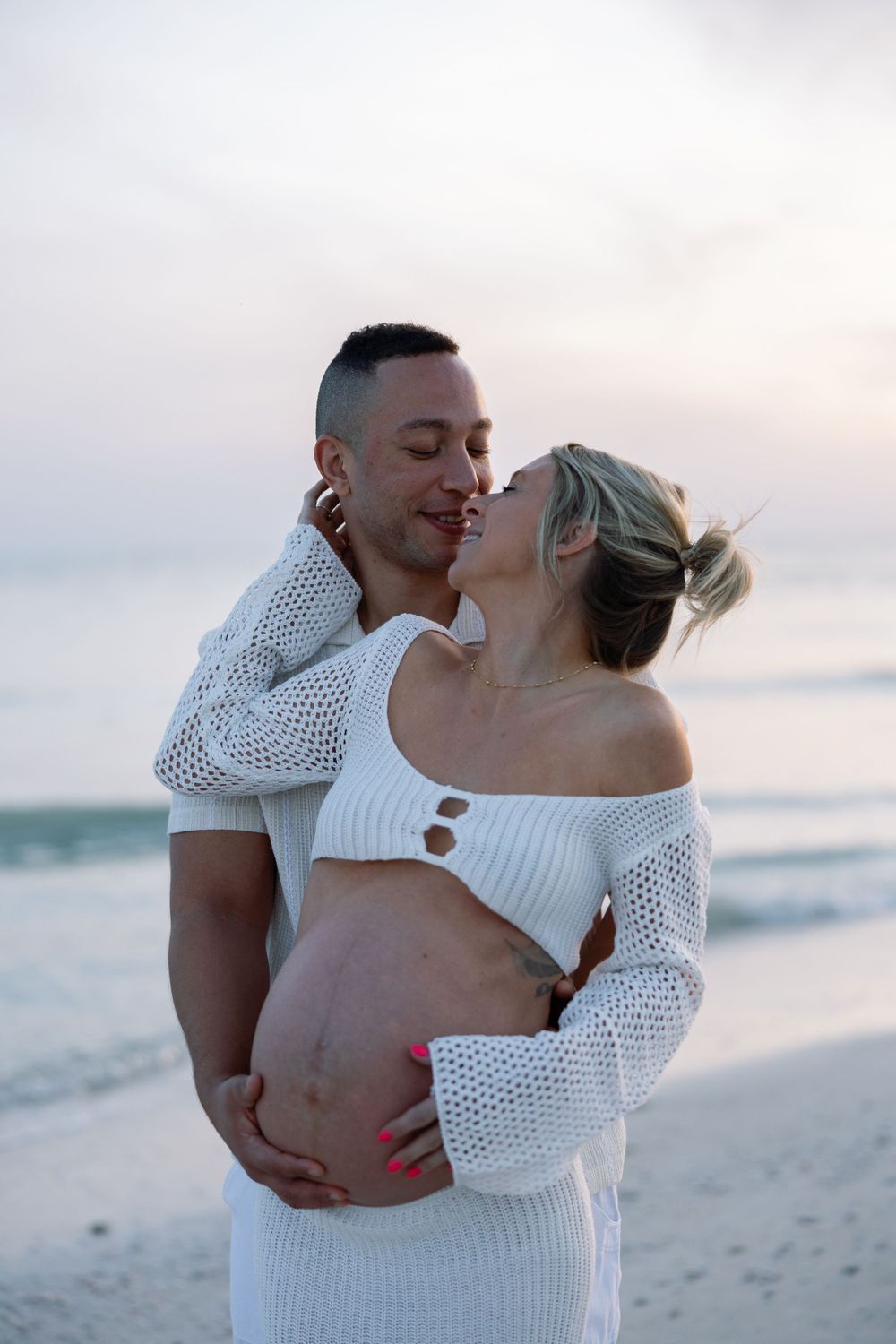 Couple shares an intimate moment on beach at sunset as they celebrate pregnancy in white knit outfits.
