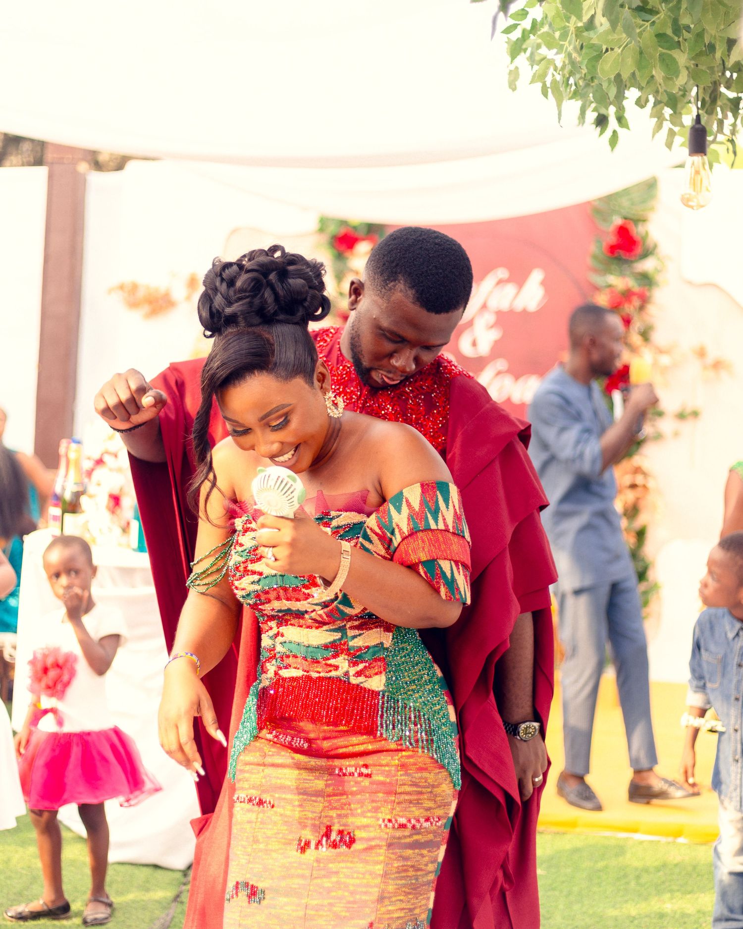 Couple in vibrant African attire share an intimate moment at an outdoor event with floral decorations.
