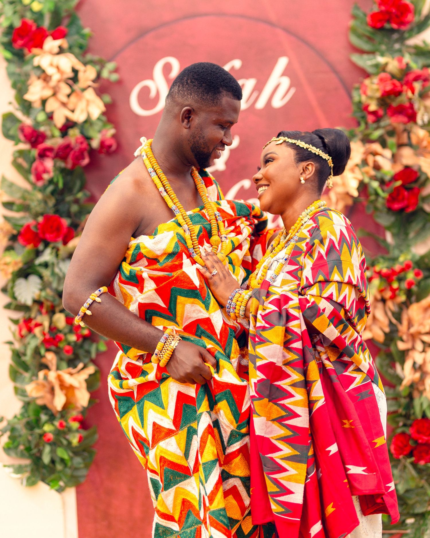 Couple in matching kente cloth poses against floral backdrop with Sefah signage.