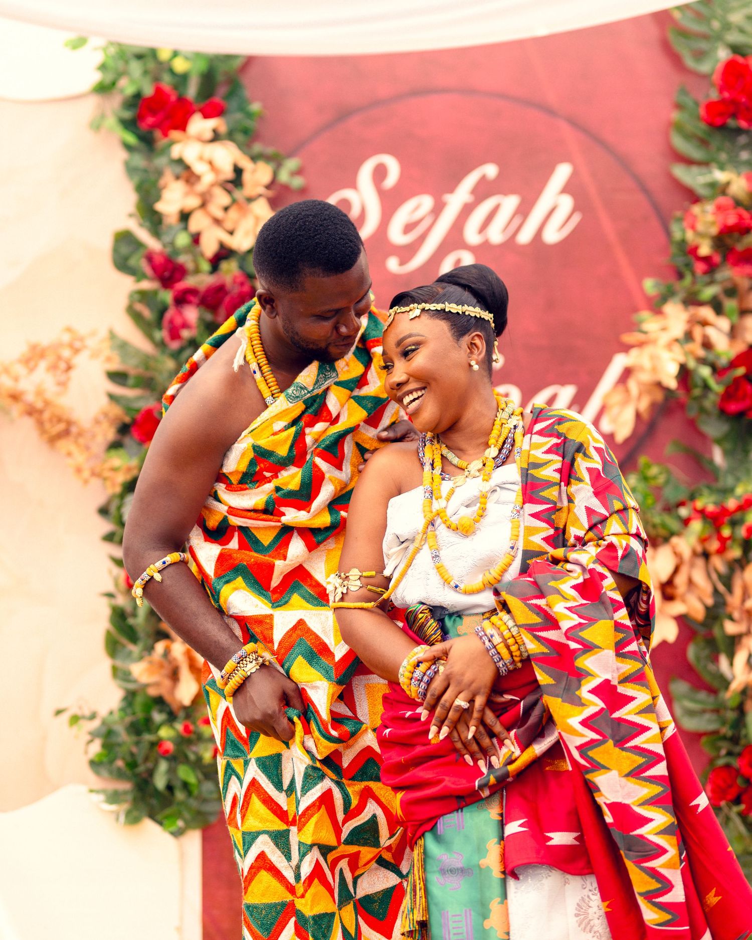 Intimate moment between couple wearing traditional kente cloth against decorative floral backdrop.