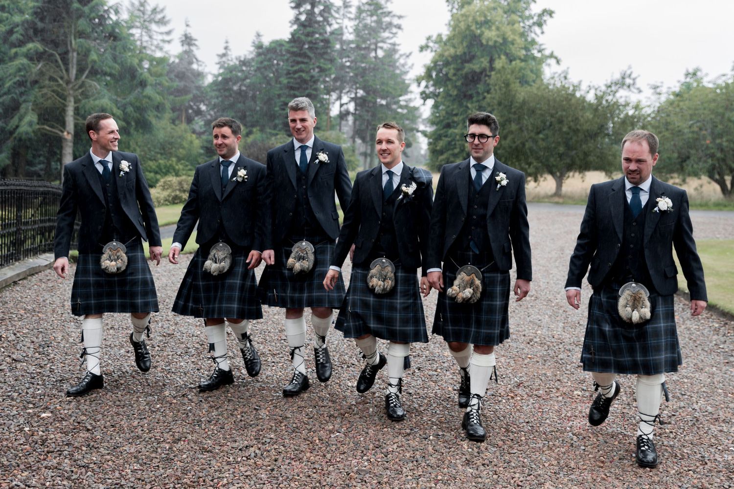 A group of six groomsmen walking together wearing traditional Scottish kilts and formal black jackets at a wedding celebration.