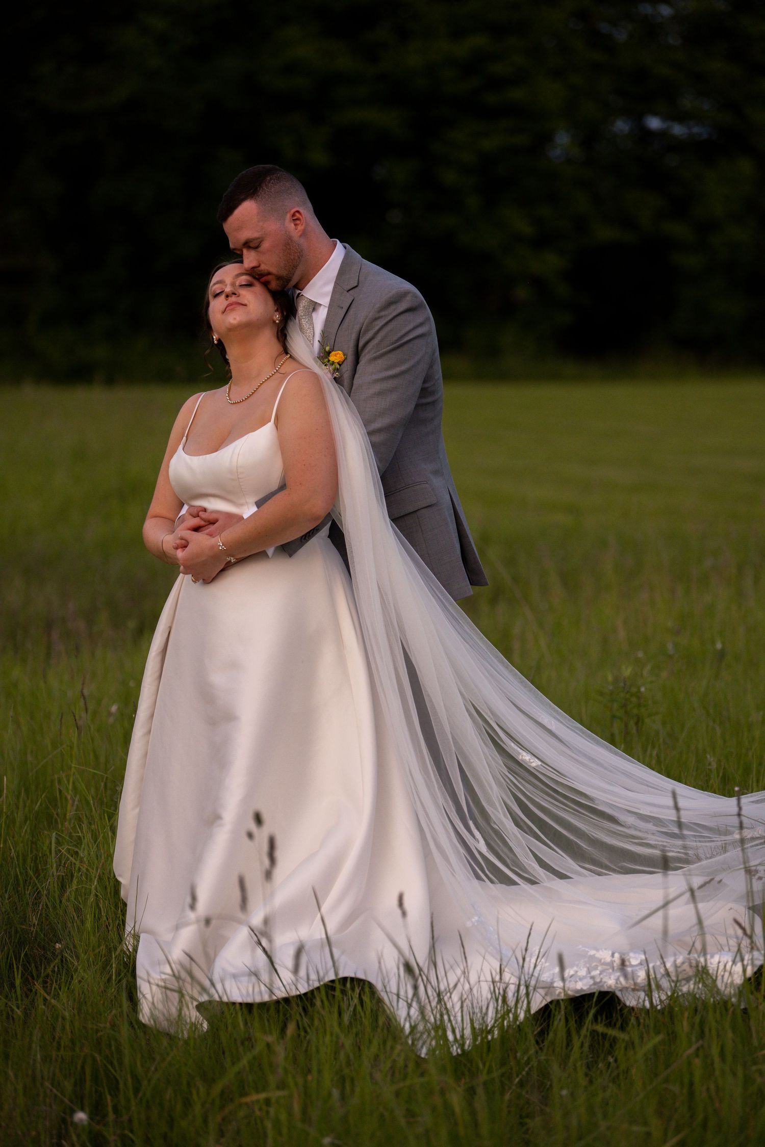 A romantic wedding couple embraces in a grassy meadow at dusk, with the bride's long veil flowing in the breeze.
