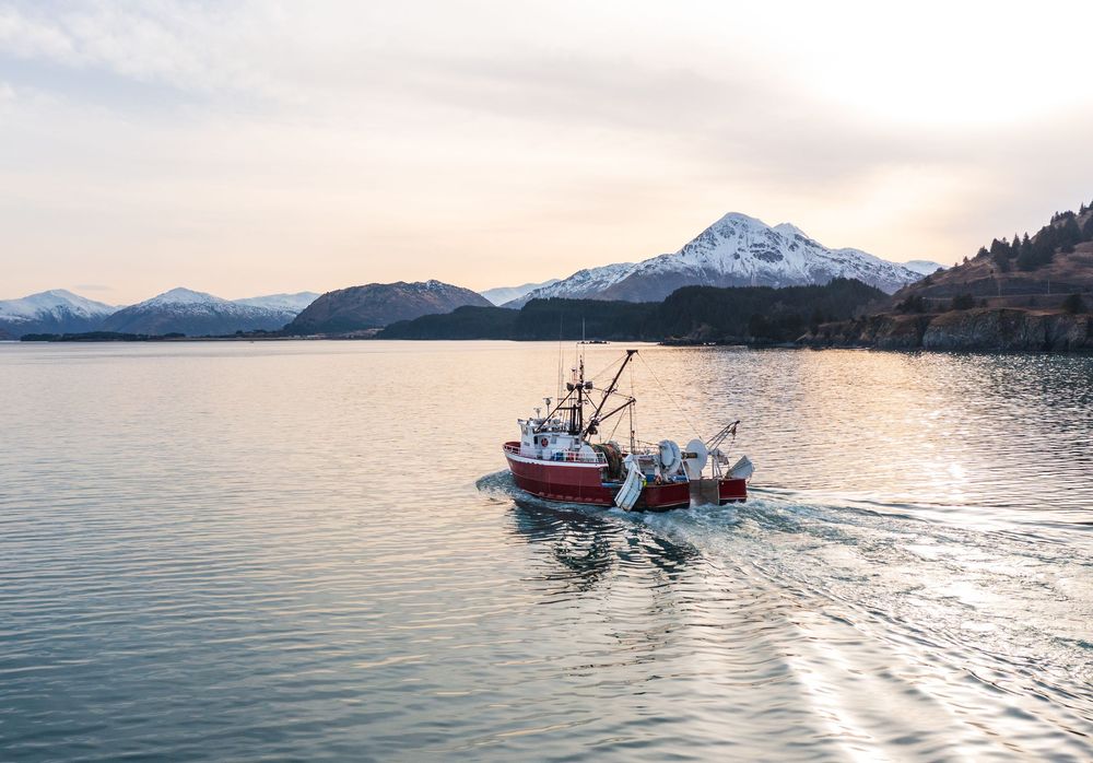 A red fishing boat cruises across calm waters with snow-capped mountains and sunset sky in the background.