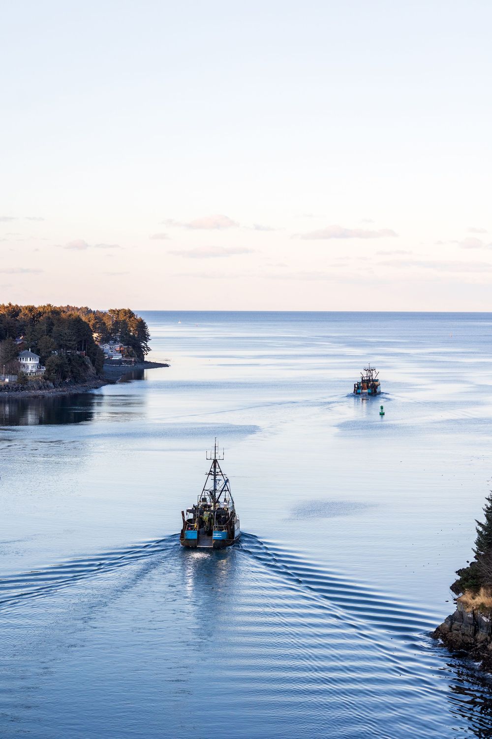Two fishing boats creating ripples in calm blue waters between rocky coastal cliffs at dusk.