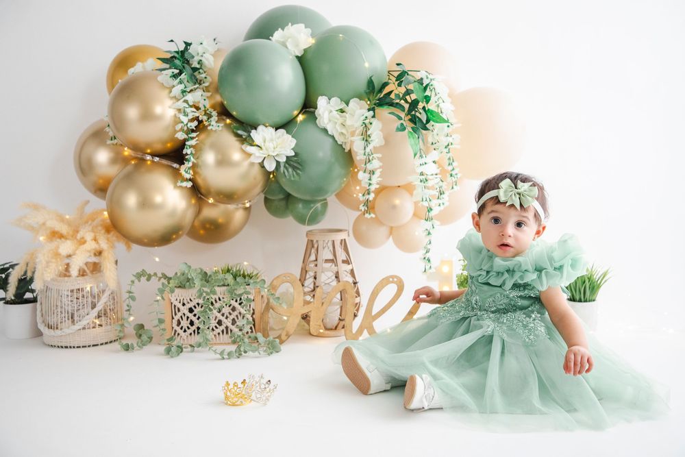 Baby girl in mint green dress sits before balloon arch with gold, sage, and white balloons at first birthday cake smash.