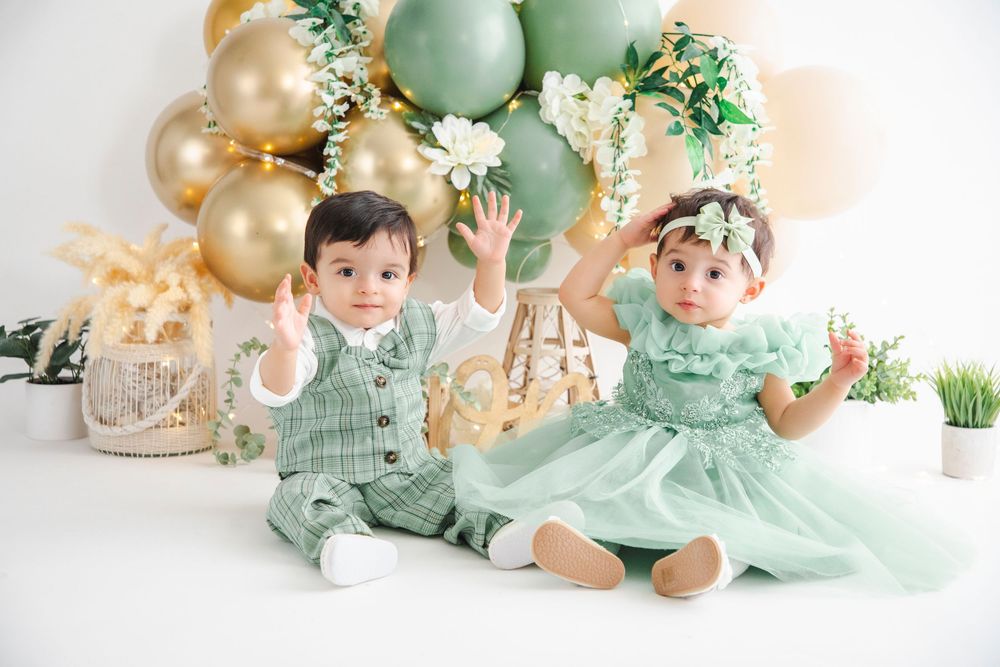 Two babies in matching green outfits sit together, waving, surrounded by gold and green balloon decorations.