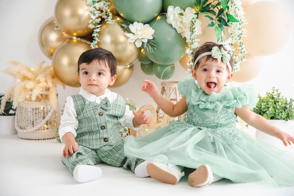Two babies in matching mint green outfits sit together surrounded by gold and green balloons at a birthday celebration.