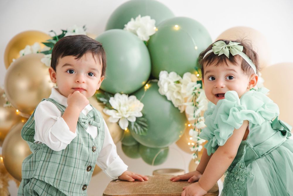 Two toddlers in matching green outfits pose with sage and gold balloons and white flowers.