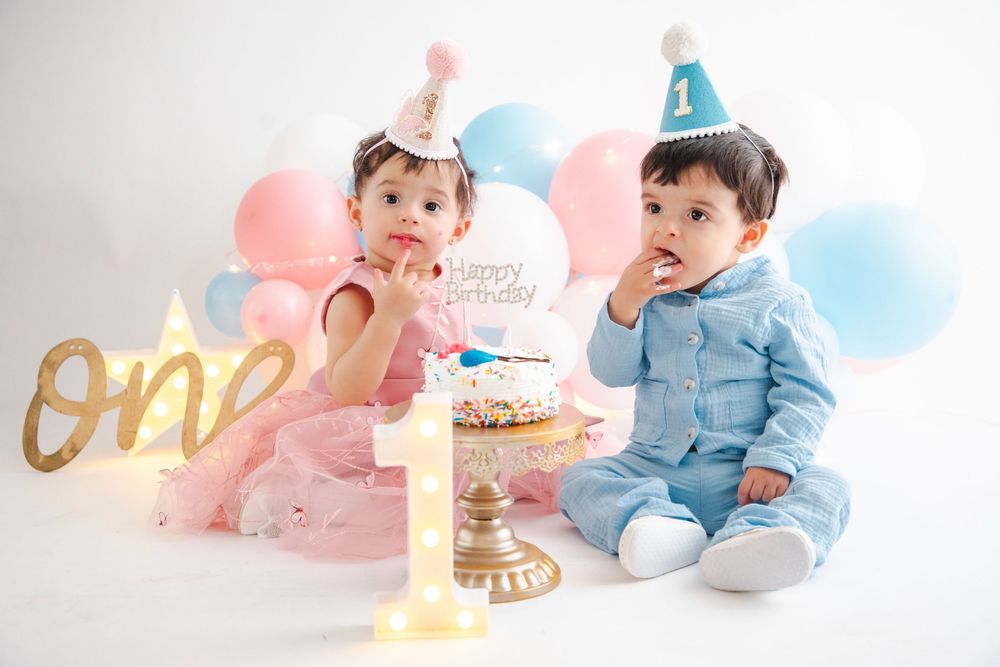 Two babies in party hats celebrate their first birthday with a smash cake surrounded by pink and blue balloons.