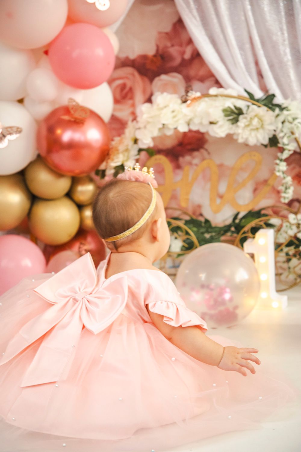 Baby girl in pink tutu dress with bow sits before first birthday balloon backdrop with floral wreath and gold \"one\" sign.