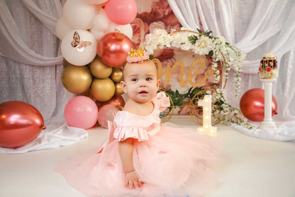 Baby girl in pink tutu dress celebrates her first birthday surrounded by rose gold and pink balloon decorations.