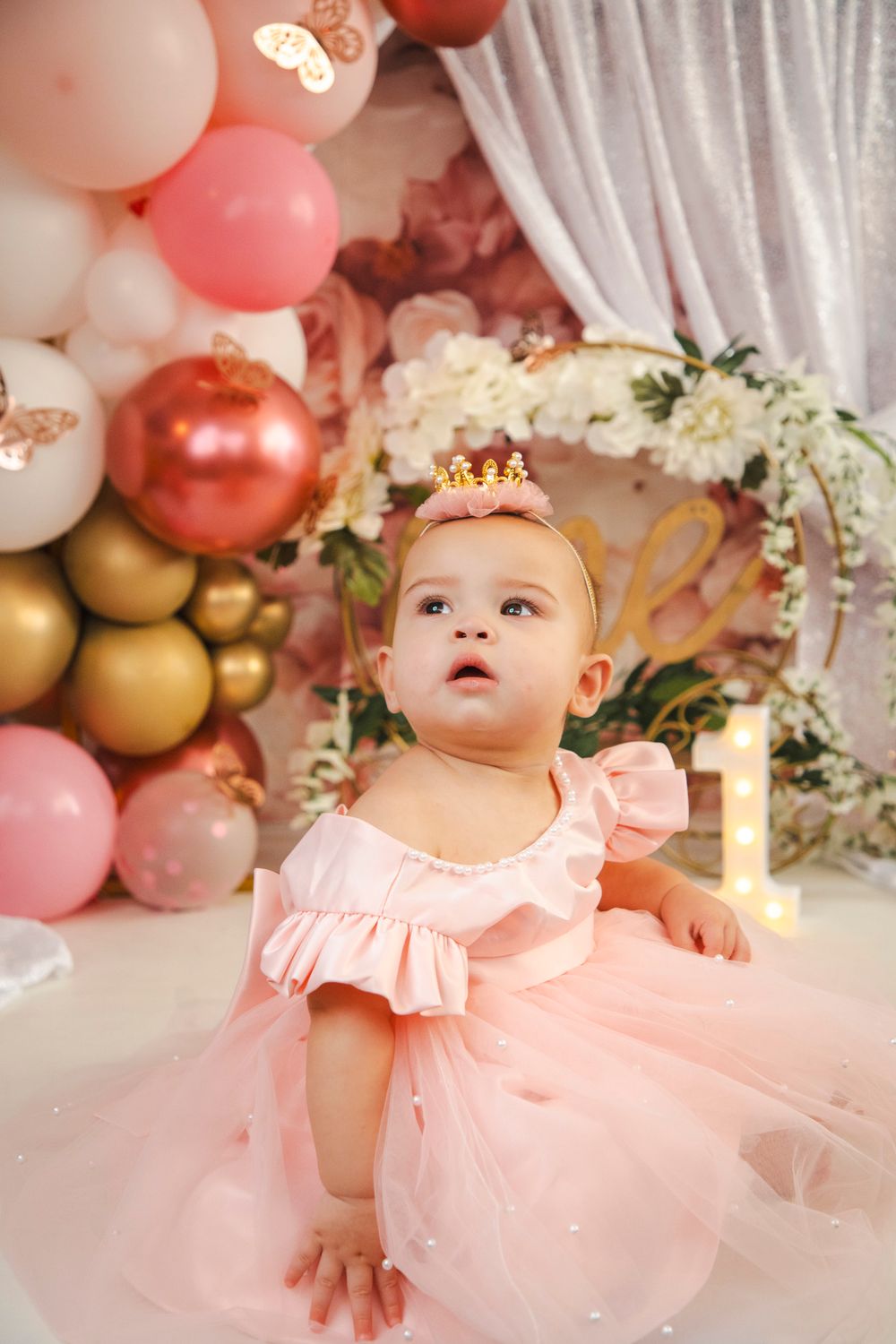 Baby girl in pink tutu dress and crown celebrates first birthday surrounded by pink and gold balloons.