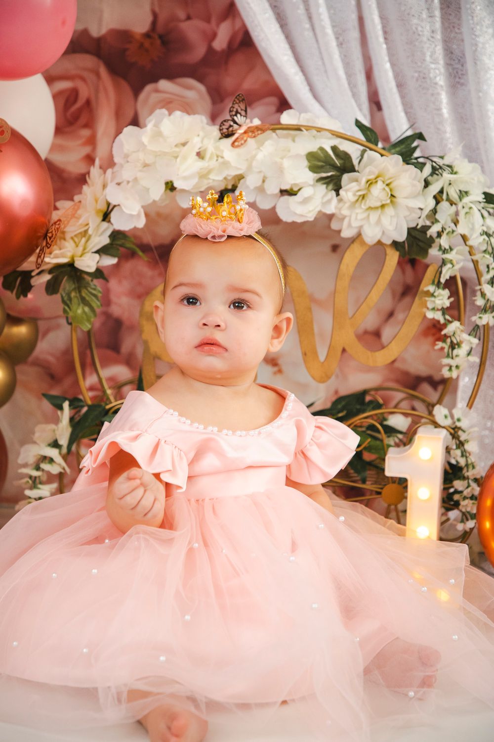 Baby girl in pink tutu dress with gold crown celebrates first birthday amid floral decor and balloons.