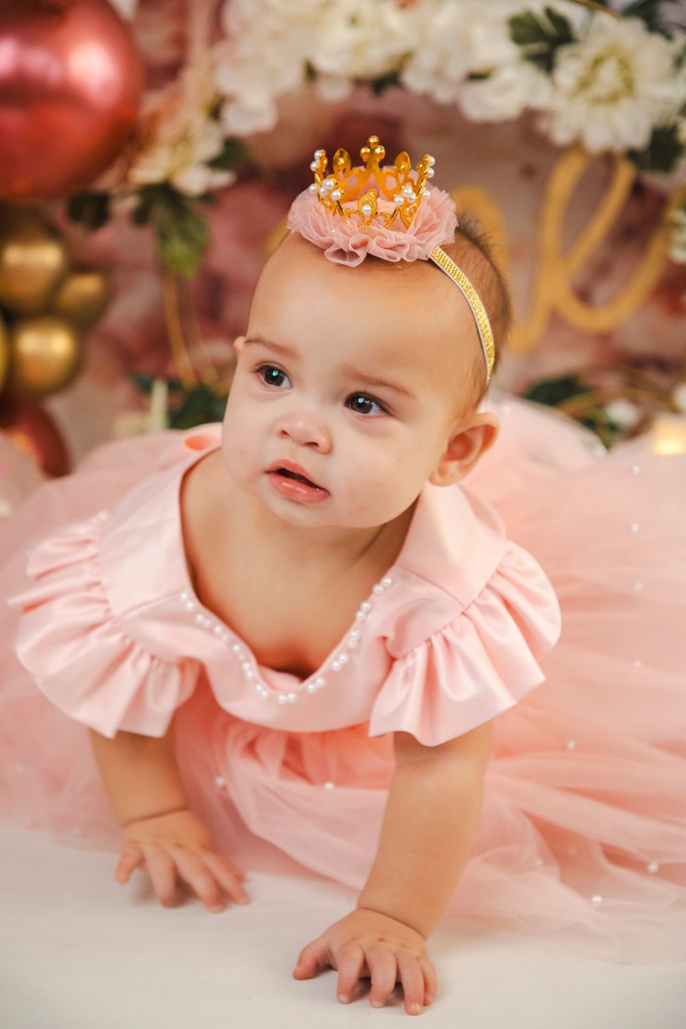 Adorable baby girl in pink tutu dress and gold crown headband crawling at her first birthday party.