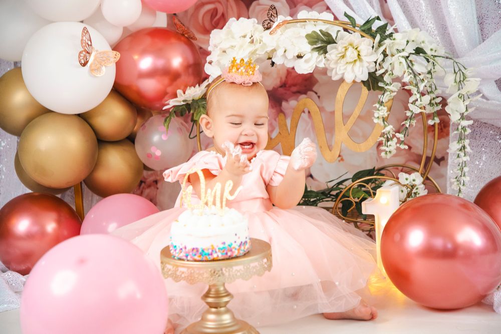 Baby girl in pink tutu smashes first birthday cake surrounded by rose gold and white balloon decorations.