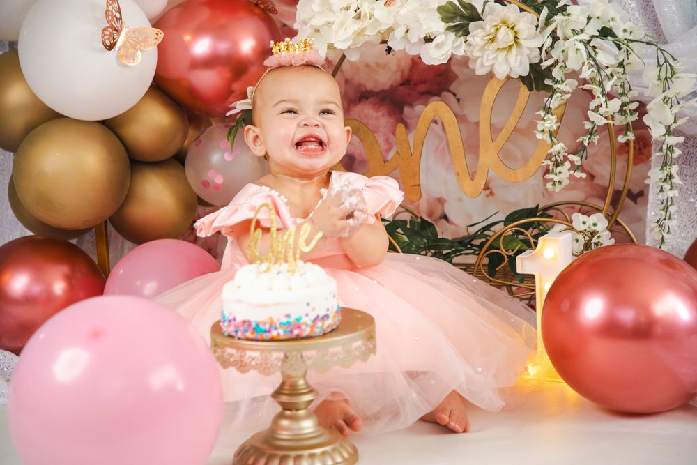 Smiling baby girl in pink tutu and gold crown celebrates first birthday with cake and balloon decorations.
